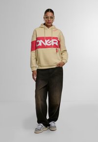 Felpa beige con striscia orizzontale rossa e lettering bianco. Pantaloni in denim scuro larghi e scarpe da ginnastica grigie con strisce bianche. Occhiali trasparenti indossati.