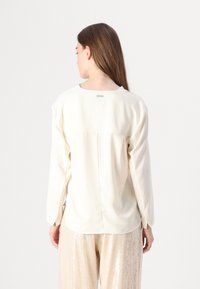 Blusa a maniche lunghe color crema con scollatura rotonda, texture liscia e cucitura centrale sul retro. Presenta un piccolo etichetta logo sulla scollatura.