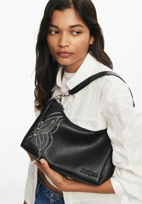 Borsa a tracolla in pelle nera con superficie testurizzata, caratterizzata da un motivo floreale ricamato bianco e dettagli metallici. Nome del marchio "Desigual" visibile.