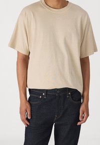 Beige bomullströja med rund halsringning och korta ärmar, med en subtil Levi's-logotyp broderad på bröstet, i kombination med mörkblå jeans.