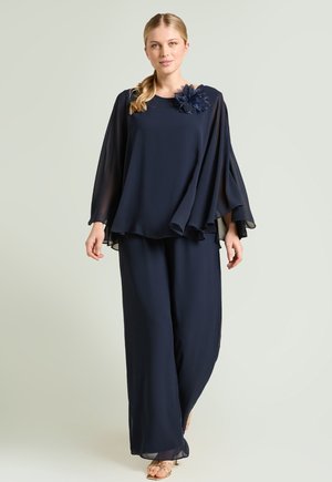 Frau trägt ein fließendes marineblaues zweiteiliges Outfit mit weit geschnittenen Hosen und einem Blütendetail an der Schulter, steht vor einem einfarbigen Hintergrund.