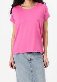 Femme portant un t-shirt rose vif à manches courtes avec des revers retroussés et une jupe en jean bleu clair sur un fond blanc uni.
