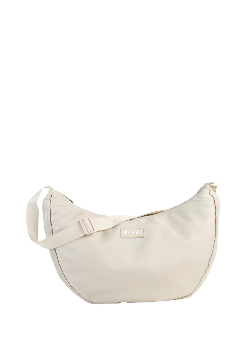 Beige stoffen schoudertas met een gebogen vorm, verstelbare band en een label met de tekst "Bench." Heeft een gladde textuur en een minimalistisch ontwerp.