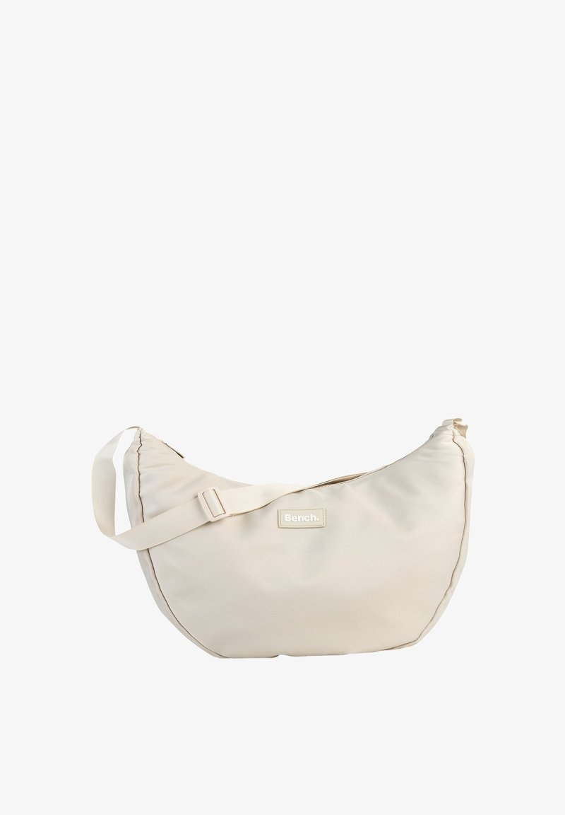 Beige stoffen schoudertas met een gebogen vorm, verstelbare band en een label met de tekst "Bench." Heeft een gladde textuur en een minimalistisch ontwerp.