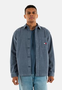 Camisa de cuadros azul con botones y un bolsillo en el pecho que lleva un logo rojo, blanco y azul. Lleva puesta una camiseta azul y jeans claros.