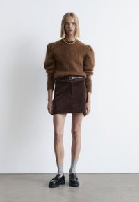 Brauner flauschiger Pullover mit puffärmel, kombiniert mit einem dunkelbraunen Cord-Minirock, schwarzen Schuhen und grauen Socken. Schlichter metallischer Gürtel als Akzent.
