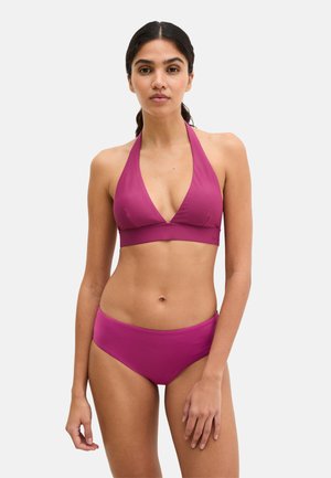 Femme aux cheveux foncés attachés en queue de cheval, portant un haut de bikini dos nu magenta et un bas assorti, debout devant un fond clair uni.
