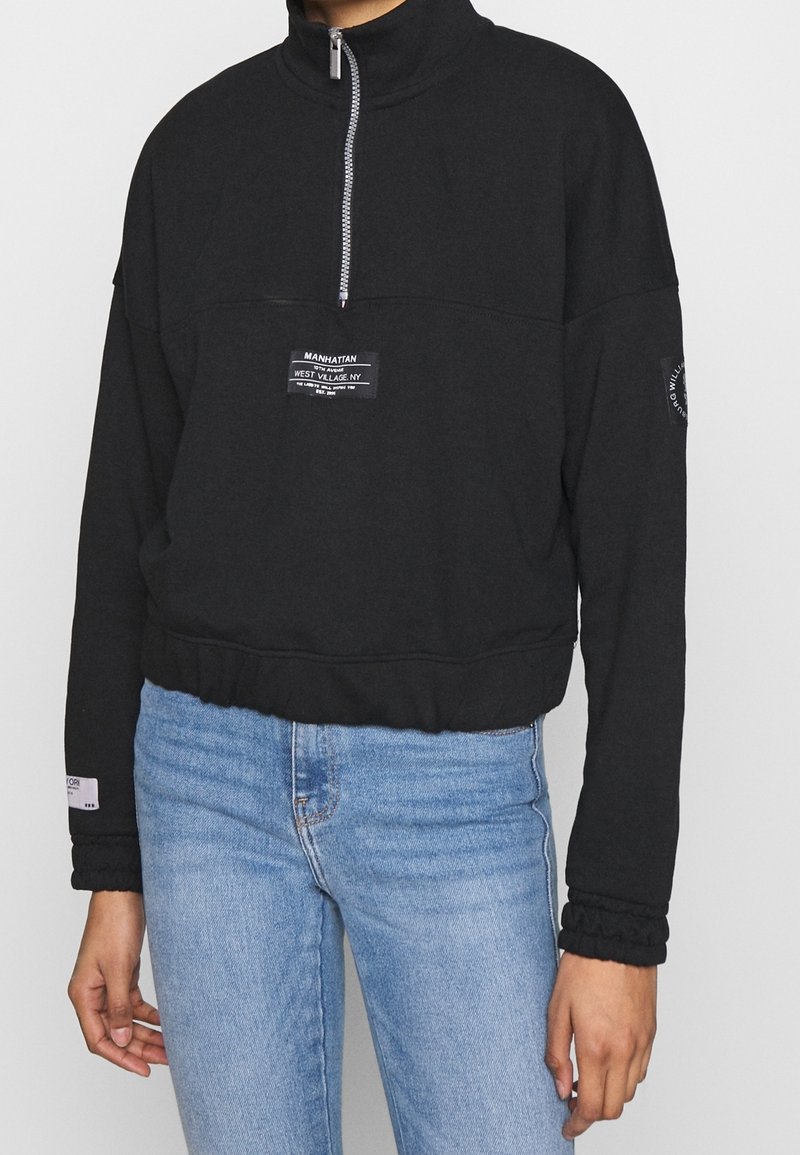 Svart sweatshirt med hög krage, delvis dragkedja, framstående patchmärke och ribbad nederkant. Har struktur i ärmsluten och en avslappnad passform.