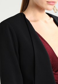 Blazer noir au design structuré et à la texture douce, associé à un haut bordeaux profond avec un motif plissé. Grande boucle d'oreille décorative visible.