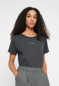 Fiatal nő hosszú, göndör hajjal, sötétszürke Pepe Jeans pólót és magas derekú szürke nadrágot visel, kezeit a zsebében tartva.