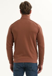 Rjava dolga zip-up jakna z visokim ovratnikom, rebrastimi manšetami in robom. Gladka tekstura z minimalnimi oblikovalskimi detajli, nosi se z modrimi kavbojkami.