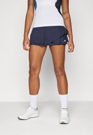 Atleet draagt witte sneakers, lange witte sokken, een marineblauwe sportrok met gelaagd ontwerp en een wit-blauw mouwloos topje.