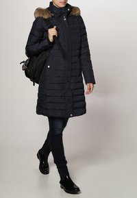 Persoon die loopt in een lange, marineblauwe gewatteerde jas met een bontgevoerde capuchon, donkere jeans, zwarte enkellaarzen, en die een zwarte rugzak op één schouder draagt.