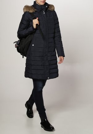 Osoba idąca w długim granatowym pikowanym płaszczu z futrzanym kapturem, ciemnych dżinsach, czarnych botkach za kostkę i niosąca czarny plecak na jednym ramieniu.