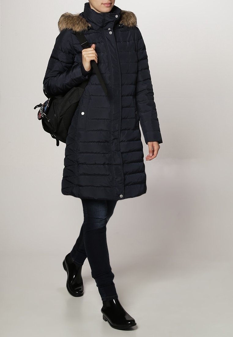Persoon die loopt in een lange, marineblauwe gewatteerde jas met een bontgevoerde capuchon, donkere jeans, zwarte enkellaarzen, en die een zwarte rugzak op één schouder draagt.