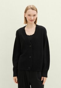 Svarte strikkede kort blazer med dyp V-hals, ribbestrikkede mansjetter og knapplukking foran. Myk tekstur, løs passform, sammen med en svart topp.
