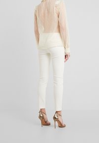 Przezroczysta, jasnobeżowa bluzka w pionowe paski z falbankowymi ramionami, zestawiona z białymi spodniami o wąskim kroju i metalicznie brązowymi sandałami na wysokim obcasie.