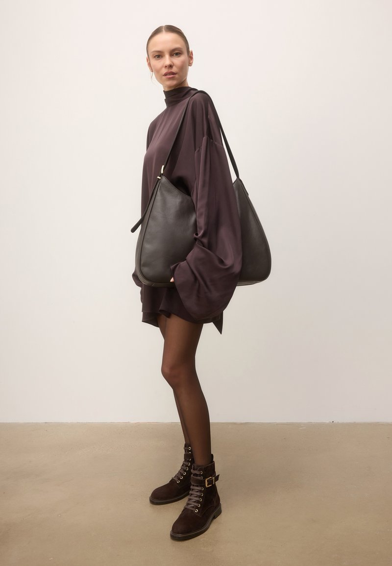 Femme portant une robe ample foncée, des collants noirs transparents, des bottes marron à lacets, portant un grand sac à bandoulière en cuir noir sur un fond uni.