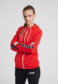 Rød zip-up hoodie med hvide snore og sorte chevron-mønstrede accenter på ærmerne, iført med sorte bukser med hvidt logo.