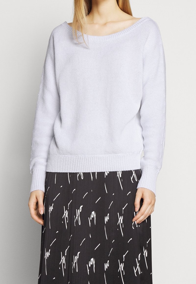 Maglione grigio chiaro a maglia con scollatura ampia e maniche lunghe, dotato di polsini e bordo a coste. Indossato sopra una gonna nera con motivo di testo bianco.