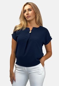 Granatowa bluzka z krótkim rękawem, z dekoltem w łódkę i detalem guzika z przodu. Gładki materiał z podwiniętymi mankietami, w zestawieniu z białymi dopasowanymi spodniami.