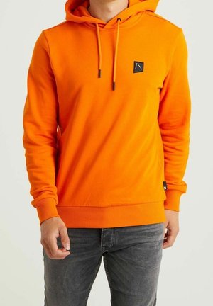 Mand iført en knaldorange hættetrøje med snore og lille sort logo på brystet, kombineret med mørkegrå jeans.