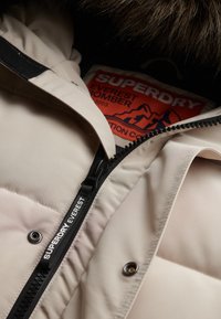 Beżowa kurtka puchowa z teksturowanej tkaniny, z czarnym zamkiem błyskawicznym z napisem "SUPERDRY EVERREST" oraz czerwoną metką z napisem "EVEREST BOMBER."