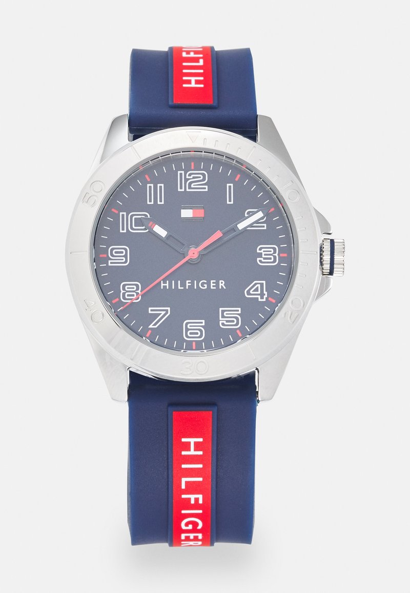 Marinblå silikonarmbandsur med rund silverfärgad boett, vit sifferdialog, röda detaljer och "HILFIGER"-logotyp. Utrustad med en roterande bezel och en boett i rostfritt stål.