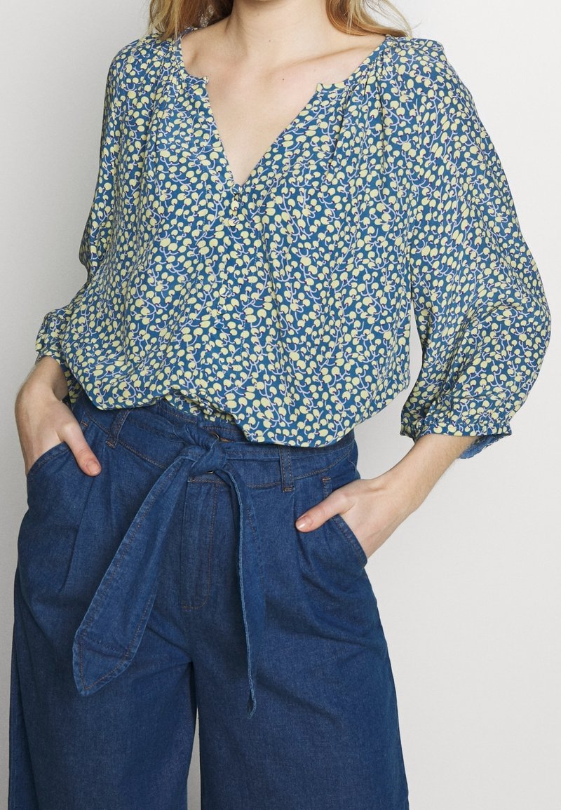 Femme portant un chemisier bleu à fleurs avec manches 3/4, rentré dans un pantalon en denim bleu taille haute avec ceinture nouée devant, mains dans les poches.
