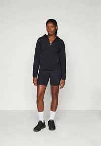 Melna zīmolota jaka ar sānu kabatām, savienota ar melnām šortiem. Modelis valkā melnus sporta apavus un baltas zeķes, stāvot uz neitrāla fona.