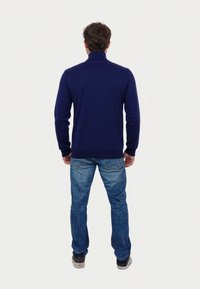 Hombre de pie de espaldas, vistiendo un suéter de cuello alto azul oscuro, jeans azul y zapatillas oscuras, sobre un fondo blanco liso.