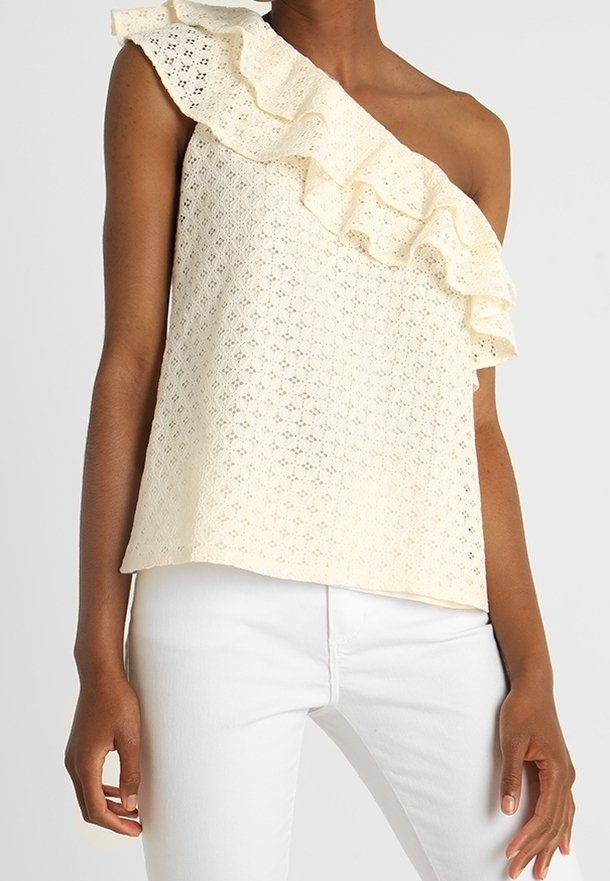 Ärmlös krämfärgad topp med ögonblommor och en volangaxel, bärs tillsammans med vita byxor på en person som står mot en enfärgad bakgrund.