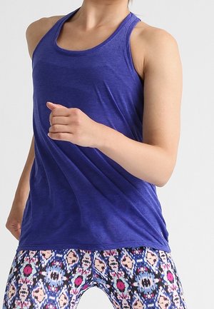 Kobieta w niebieskiej, bezrękawnikowej bluzce i kolorowych legginsach z wzorem, pozująca jak podczas biegu lub ćwiczeń na jednolitym tle.