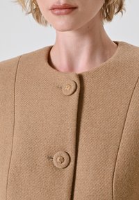 Cappotto beige con un tessuto testurizzato, scollatura rotonda e due bottoni, uno con un accento dorato vicino all'area del petto.