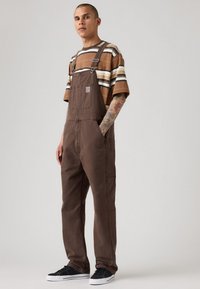 Man in een bruine overall met een voorzak, een gestreept bruin-wit T-shirt met korte mouwen, zwarte sneakers en zichtbaar armtattoo, staande.