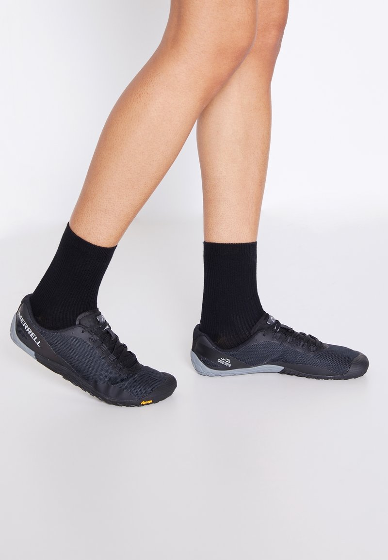 Gambe con calzini neri alla caviglia e scarpe Merrell Vibram in stile barefoot nere, in piedi su uno sfondo chiaro e semplice.