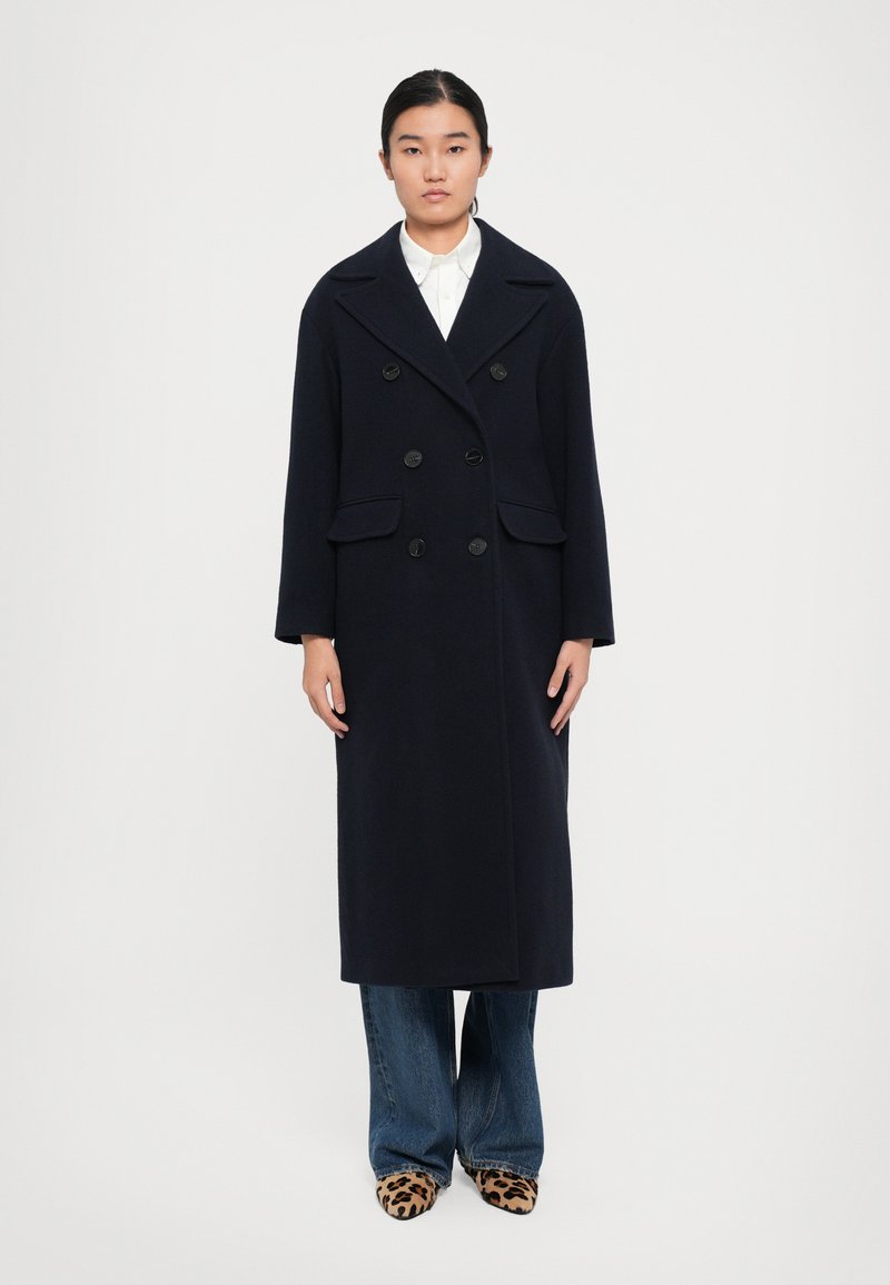 Donna in piedi con indosso un lungo cappotto doppiopetto blu navy, camicia bianca con colletto, jeans blu e scarpe a punta con stampa leopardata, di fronte a uno sfondo neutro.