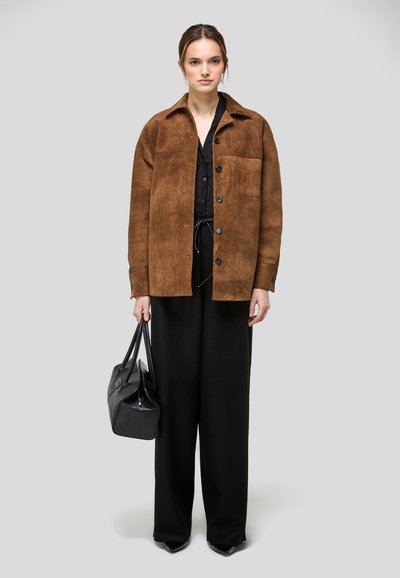 Nő barna, gombos dzsekit visel fekete ruha felett, fekete táskát tart, miközben egy egyszínű szürke háttér előtt áll.