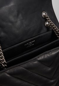 Černá kožená kabelka s prošívaným vzorem, stříbrným řetízkovým popruhem a vnitřní značkovou nálepkou s nápisem „Kurt Geiger, I am leather."