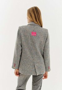 Blazer s kostkovaným vzorem v šedé barvě s multicolor fleky, má štítek na zadní straně a knoflíky v růžové, žluté a fialové barvě na manžetách.