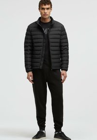 Black puffer jacket with horizontal quilted design, high collar, and zip closure, worn over a black shirt and dark pants, paired with black sneakers.