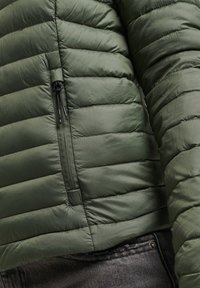 Grön vadderad jacka med vertikal dragkedjeförsedd bröstficka, bärs över mörka denimjeans, visar midja och arm.