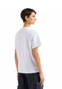 Hvid bomulds T-shirt med rund halsudskæring og korte ærmer, der har en afslappet pasform og glat tekstur. Båret med mørke bukser, set fra ryggen.