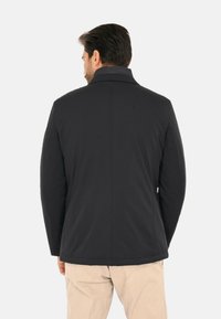 Giacca nera da uomo con collo alto, tessuto liscio, taglio sartoriale e cuciture minime. Nessuna ferramenta o motivo visibile.