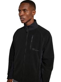 Mand iført en sort fleecejakke med lynlås, med en brystlomme med lynlås og lille trykt tekst på venstre side.