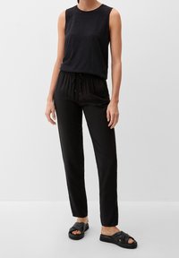 Top noir sans manches avec des détails texturés aux épaules, associé à un pantalon noir ample à cordon de serrage et des sandales plates noires à larges brides.