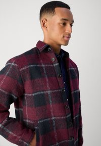 Veste en laine à carreaux bordeaux pour homme avec des accents noirs et gris, fermeture à bouton et deux poches latérales. Texture douce et coupe décontractée.