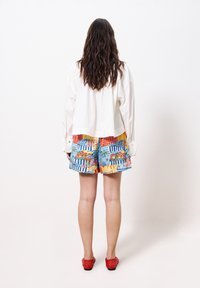 Weißes kurzes Hemd, farbenfrohe gemusterte Shorts mit blauen, roten, gelben und grünen Akzenten sowie roten Slippers. Glatte Textur, lässiger Stil.