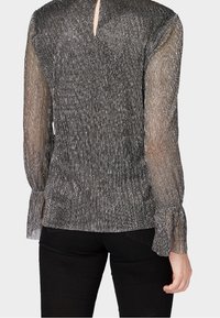 Metallic strukturierte Bluse mit durchsichtigen langen Ärmeln und Rüschenmanschetten, getragen mit schwarzen Hosen aus der Rückenansicht.