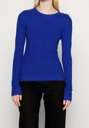 Femme portant un pull côtelé bleu roi à manches longues avec des détails de boutons sur les épaules et un pantalon noir sur un fond uni.